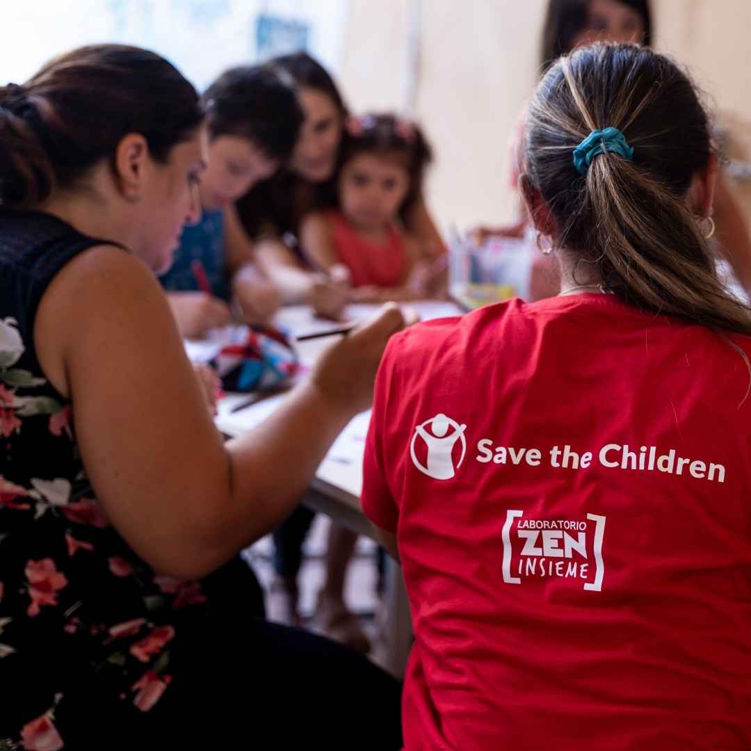 Operatrice di Save the Children in maglia rossa, seduta a un tavolo, mentre segue un'attività di sostegno scolastico a bambini.